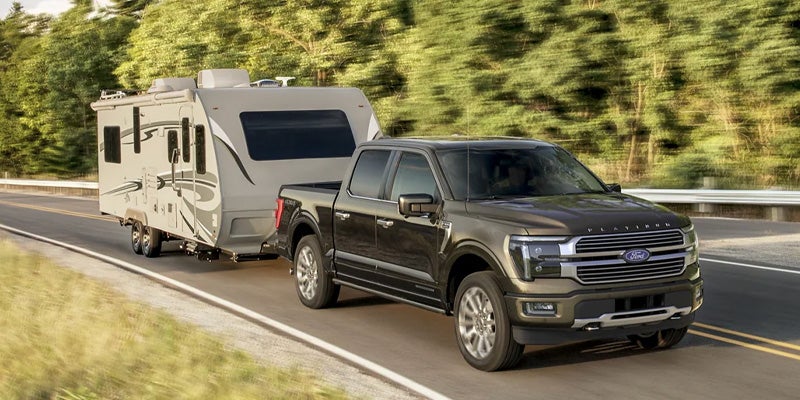 Dark brown Ford F-150 Platinum truck towing a light gray travel trailer on a road with blurred green trees.