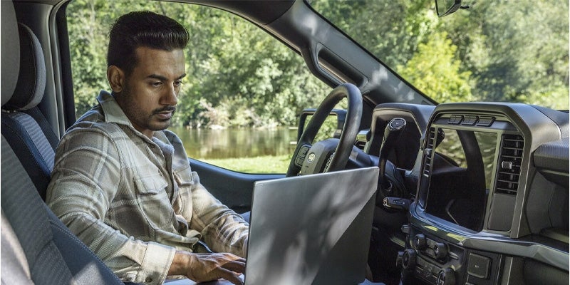 Man in a truck’s driver seat using a laptop on a flat console, with trees and water visible outside.