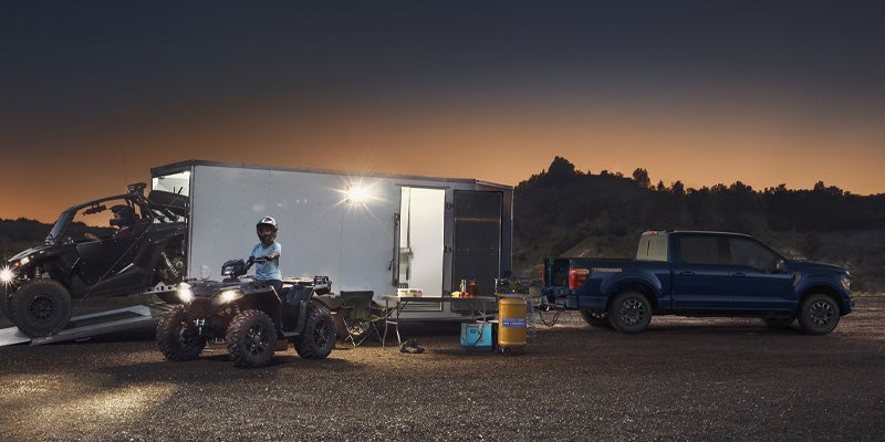Blue pickup truck towing a trailer with ATVs at dusk, camping gear set up, and a child on an ATV.