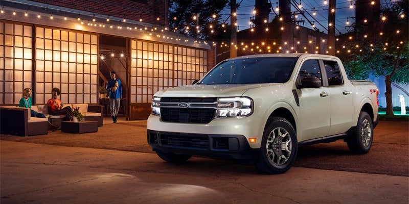 Light-colored Ford Maverick truck parked at night in front of a building with string lights and people socializing.