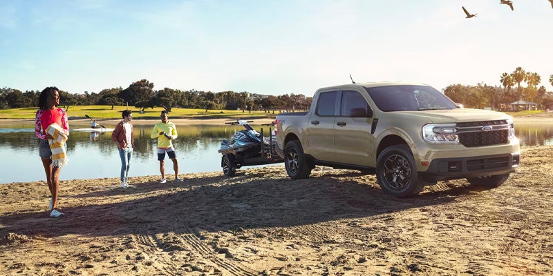 Tan Ford Maverick truck towing a jet ski on a trailer at a sandy lake shore with three people.