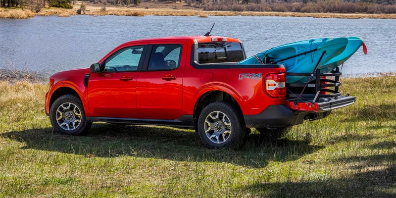 Red pickup truck with a blue kayak in its bed, parked on grassy shore next to a lake.