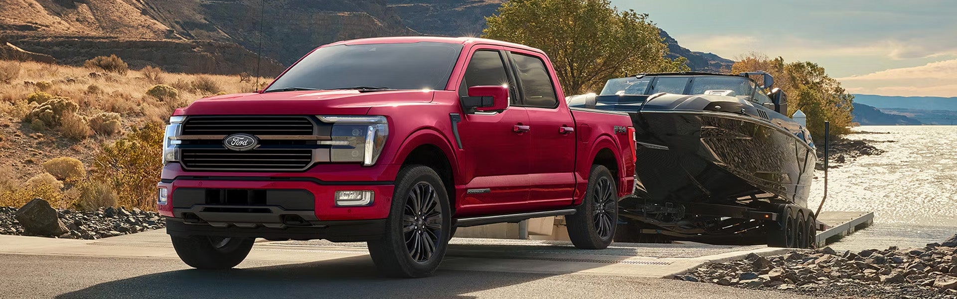 Red Ford F-150 truck towing a black boat down a concrete ramp into a lake, with mountains in the background.