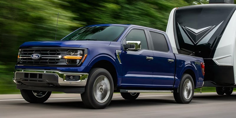 Blue Ford F-150 pickup truck towing a large white and black RV on a road with blurred green background.