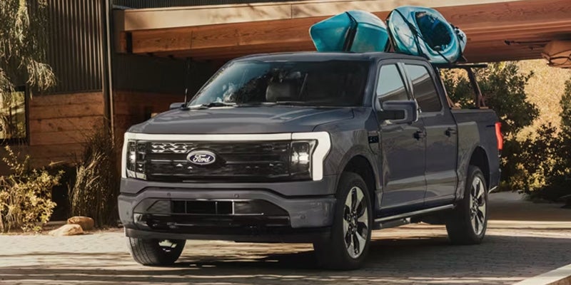 Dark gray Ford F-150 Lightning pickup truck with two blue kayaks on a bed rack, parked in front of a modern home.