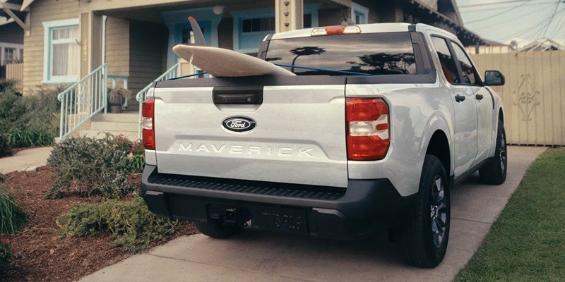 Rear view of a silver Ford Maverick pickup truck with a surfboard in its bed, parked on a driveway in front of a house.